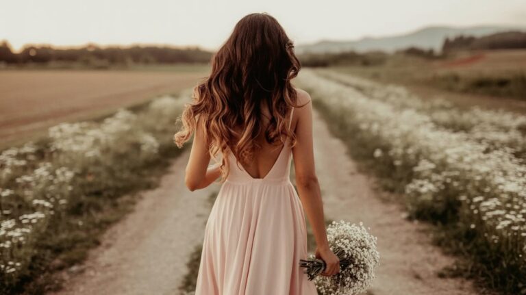 A woman with long, wavy hair wearing a backless pink dress walks down a flower-lined dirt path, bouquet in hand—an idyllic moment perfect for a faith lifestyle blog, with fields and mountains stretching behind her.